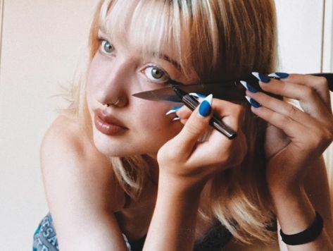 blonde, young girl doing her eyeliner with a knife placed on the side of her eye to help line up the make up. she uses an eyeliner pen and the knife as a ruler against her skin. the photo is edited to look like it was taken off a film camera. Drawing Eyeliner With Knife, Doing Eyeliner With Knife, Knife To Neck Pose, Woman Holding Knife Reference, Girl Holding Knife Reference, Man Holding Knife Reference, Person With Knife Pose, Person With Knife Reference, Person With Knife Pose Reference