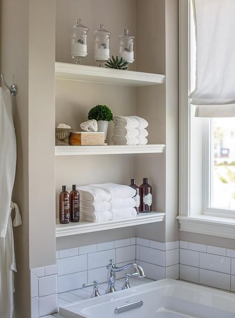 White stacked floating shelves are mounted in a nook above a drop in bathtub fitted with a white subway tiled deck holding a polished nickel vintage hook and spout tub filler in front of white subway backsplash tiles fixed beneath a window dressed in a white roman shade accenting gray wall paint. Bathroom Shelves Above Tub, Shelving Above Tub, Alcove Tub With Half Wall, Over Tub Shelves, Shelving Over Bathtub, Shelves Above Tub, Bathtub Shelf Ideas, Window Above Bathtub, Shelves Above Bathtub