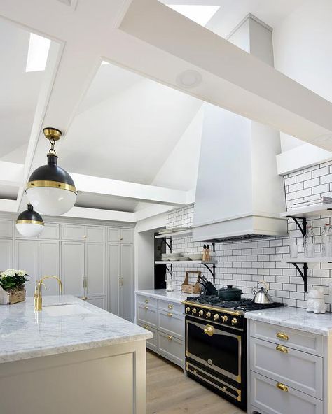 Illuminated by skylights fitted to a sloped ceiling, this gorgeous gray and white kitchen boasts a white hood mounted to white subway backsplash tiles accented with black grout above a La Cornue CornuFe Range and between iron and marble shelves. Range Hood On Sloped Ceiling, Range Hood With Vaulted Ceiling, Patagonia Kitchen, Range Hood Vaulted Ceiling, Subway Tiles Kitchen, Kitchen Cabinets Painted Grey, Black And White Backsplash, Marble Shelves, Grey Painted Kitchen