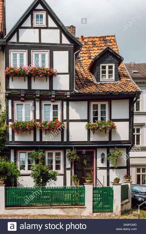 View of a traditional old German house with timber framing, windows decorated with red flowers and a green wooden fence in front of it, Quedlinburg. Stock Photo German Exterior House, German Houses Traditional, Traditional German House, German Tudor House, German House Aesthetic, German Architecture Traditional, Sims 4 German House, German Home Exterior, German Townhouse