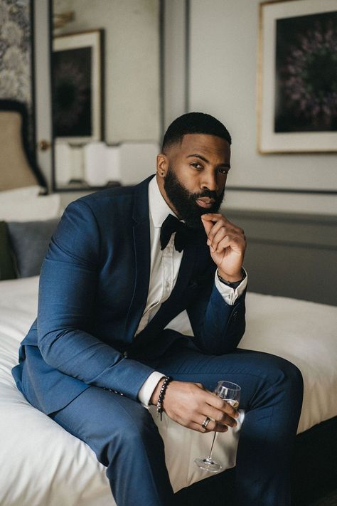 Black bearded groom in a navy blue suit with black bow tie sitting on the bed on the wedding morning Wedding Suits Men Black Groom Attire Navy Blue, Navy Blue Suit Black Lapel Wedding, Midnight Blue Tuxedo Wedding Groomsmen, Bow Tie Men's Outfit, Groom Navy Tux Groomsmen Black, Groom Black Bow Tie Tuxedo, Black Bride And Groom, Dark Navy Suit, Navy Blue Groom