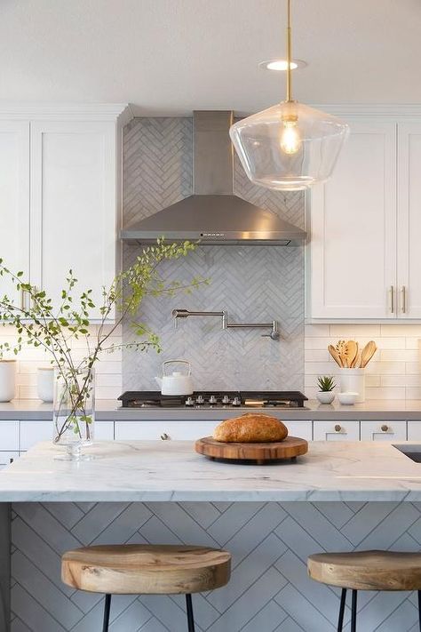 White shaker cabinets flank a stainless steel vent hood fixed to gray herringbone pattern tiles over a satin nickel swing arm pot filler fitted above a gas integrated cooktop. Kitchen Backsplash Herringbone, Backsplash Herringbone, Kitchen Vent Hood, Replacing Kitchen Countertops, Kitchen Vent, White Shaker Cabinets, Kitchen Backsplash Designs, Pot Filler, Vent Hood