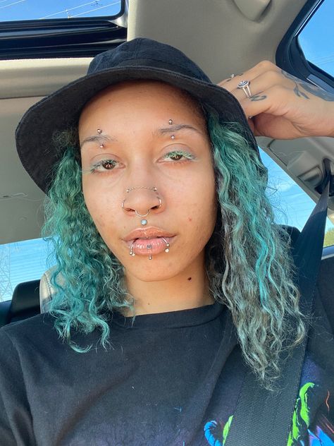 Picture of a person with curly hair displaying their piercings. They have an silver curved barbell in the center of each eyebrow. They have double opal high nostril piercings, each sitting above a nostril ring. They have a septum that hangs in the center of they’re nose just above an opal stud in the dip of the top lip. They also have a labret in the center of they’re bottom lip framed by a ring on each side of the bottom lip. Snakebites And Labret, Double Vertical Eyebrow Piercing, Labret And Snakebites, Eyebrow Piercing Double, Centered Eyebrow Piercing, Snakebites And Vertical Labret, Double Brow Piercing, Hoop Eyebrow Piercing, Vertical Labret And Snakebites