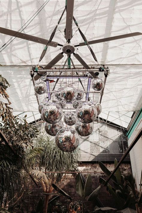 An installation of tiered disco balls risen above the aisle in a lush greenhouse filled with green plants and trees at an alternative venue in Los Angeles. Disco Ceiling, Lush Greenhouse, Eclectic Diy, Groovy Wedding, Paradise Plant, 70s Decor, Flower Installation, Shimmer Lights, Ceiling Installation