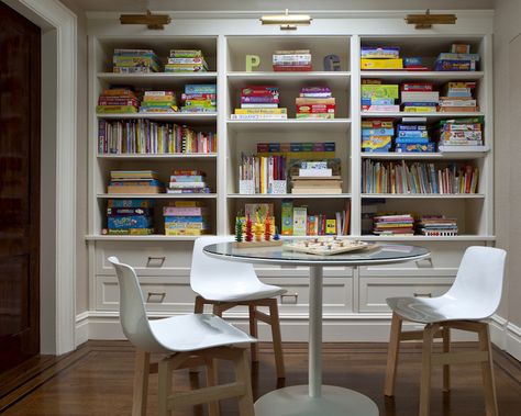 Fantastic game room features wall of built-in bookshelves filled with board games and books illuminated by antique brass picture lights situated behind glass-top game table surrounded by white plastic chairs White Plastic Chairs, Board Game Room, Garage Game Rooms, Homework Room, Basement Games, Game Room Kids, Board Game Storage, Game Room Basement, Game Storage