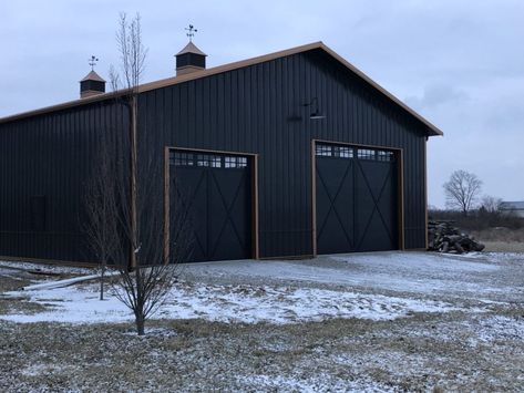 This 50' by 72' machine storage and garage was built with black steel panels and a bronze trim. The bronze roof and dual cupolas with weather vanes on top of the building add a distinguished final look. 80x100 Metal Building, Black Metal Building With Wood Accents, Black Workshop Exterior, Black And Copper Metal Building, Black Steel Roof House, Black Outbuilding, Black Morton Building, Black Steel Building, Steel Building Color Combinations