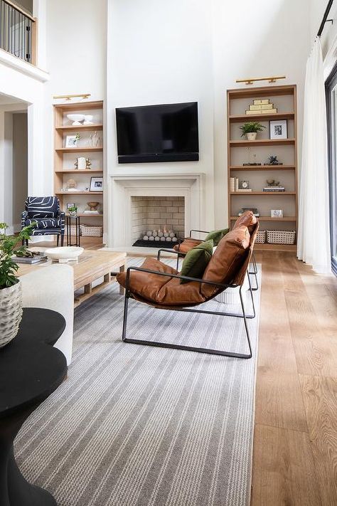 Spacious living room features brown leather accent chairs accented with forest green velvet pillows, a tan oak coffee table atop a gray stripe rug, and a TV mounted over a white fireplace flanked by oak built in shelves lit by long brass lights. Leather Brown Chair Living Room, Brown Leather Arm Chair Living Room, White Sofa With Brown Accent Chairs, Velvet Living Room Chairs, Green White Oak Living Room, White Couch Brown Leather Chair, Tan Leather Armchair Living Rooms, Feature Chairs Living Room, Tan Leather Accent Chair Living Rooms