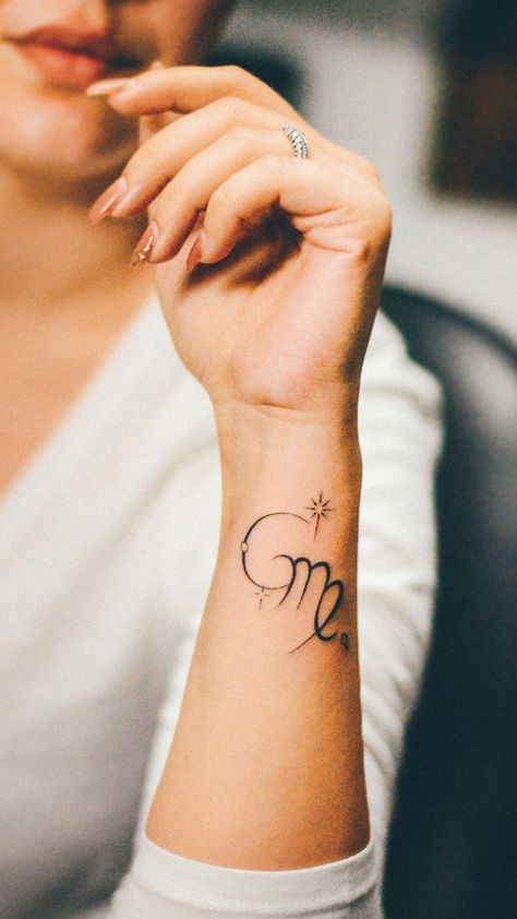 A woman's forearm is adorned with a delicate tattoo of the Virgo zodiac sign. The design features a stylized 'M' with a loop, representing the maiden, and is embellished with a small heart and twinkling stars, suggesting a personal connection to the celestial theme. Virgo Women Tattoo, Virgo Tattoo Ideas, Virgo Sign Tattoo, Virgo Tattoo Designs, Virgo Symbol, Zodiac Sign Virgo, Orange Nail Designs, Virgo Tattoo, Crescent Moon Tattoo