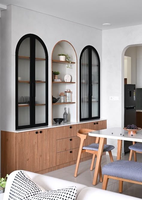 stylish crockery unit with arched niches, echoing the home's curvaceous theme. A central open shelf with wooden beading is flanked by cabinets featuring ribbed glass doors. To emulate a natural rock niche, we textured the upper storage units with raw concrete paint. Below, three teak-finished cabinets offer practical storage, keeping the space clutter-free. Crockery Units Modern, Crockery Unit Design Dining Rooms, Crockery Cabinet Design, Wall Niches, Crockery Cabinet, Crockery Unit Design, Crockery Design, Concrete Paint, Glass Cabinets