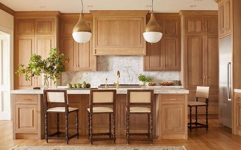 Lit by brass pendants, a honey stained oak kitchen island is accented with cabinets adorned with brass pulls topped with thick white marble fitted with a sink and polished brass gooseneck faucet lined with wood barstools upholstered in cream fabric. White Oak Kitchen Cabinets, Natural Wood Kitchen Cabinets, Honey Oak Cabinets, White Oak Kitchen, Natural Wood Kitchen, Oak Kitchen Cabinets, Honey Oak, Wood Kitchen Cabinets, Oak Kitchen