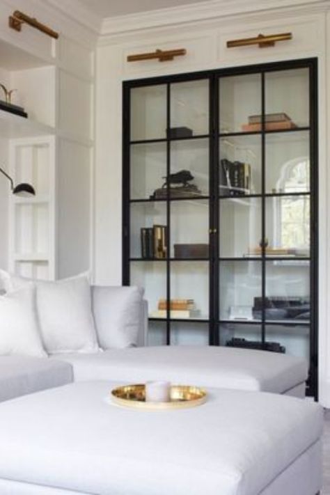 We greatly appreciate a beautiful built-in featuring glass doors! We also love the look of these black-frame glass doors and their contrast to this bright, white living area. Read our blog post for more built-in design inspiration. #interiordesign #interiordesigner #built-ins #built-in #shelves #glassdoors #whitesofa #sofa #shelfstyling #gallerylights #bookshelves Glass Shelves In Bathroom, Glass Shelves Decor, Glass Cabinet Doors, Hus Inspiration, Glass Cabinet, Book Shelf, Cheap Home Decor, Glass Shelves, Built Ins