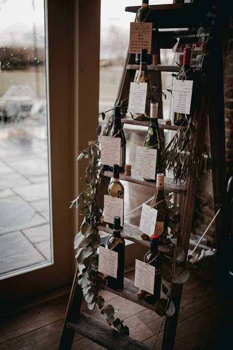 Wooden ladder seating chart finished with empty wine bottles and green foliage decor | The Vedrines Unique Seating Chart Wedding Display, Wedding Sitting Charts, Wine Cork Seating Chart, Blanket Ladder Seating Chart, Bottle Seating Chart Wedding, Wine Bottle Table Plan, Wine Seating Chart, Wedding Wine Bottle Decor, Wine Seating Chart Wedding