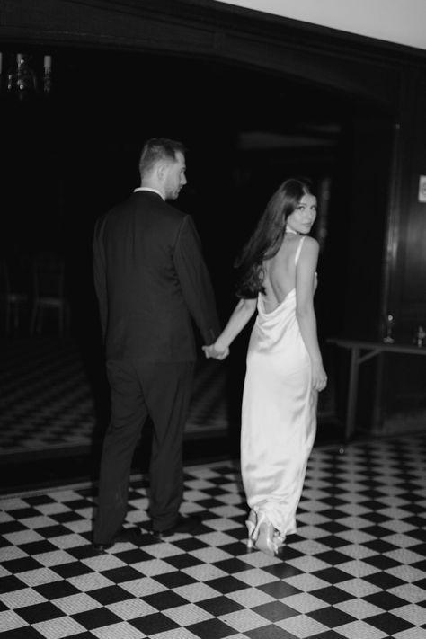 Couple standing on black and white tile floor. Women in white dress looking back at camera, man in black tux looking forward Black White Engagement Photo, Chic Engagement Shoot, Dressed Up Engagement Photos, Sophisticated Engagement Photos, Hollywood Glam Engagement Photos, Hotel Bar Engagement Photos, Old Money Aesthetic Engagement Photos, Cool Girl Engagement Shoot, Black Tie Engagement Photos