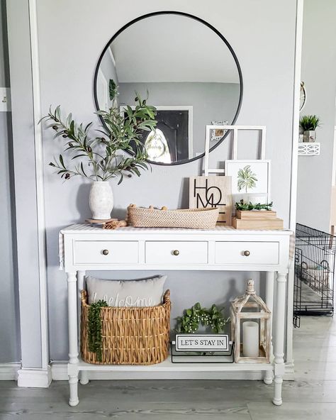 A black steel-framed mirror hangs over a white console table with a neutral runner, woven storage baskets, and a stone vase with olive stems. A wooden home sign leans against a white window frame. A wood lantern sits on the lower shelf next to a wreath and lettered sign.