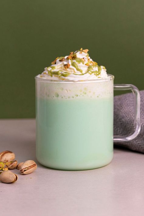 A green coloured pistachio and rose flavoured hot chocolate, made with white chocolate powder. The drink is in a transparent mug, sat on a grey table next to some pistachios, all against a dark green background. The drink is topped with whipped cream, pistachio flavoured sauce and crushed pistachios. Rose Hot Chocolate, Pistachio Hot Chocolate, Romantic Drinks, Pistachio Syrup, White Hot Chocolate Recipe, Chocolate Coffee Beans, Milkshake Machine, Coffee Syrups, Slushie Recipe