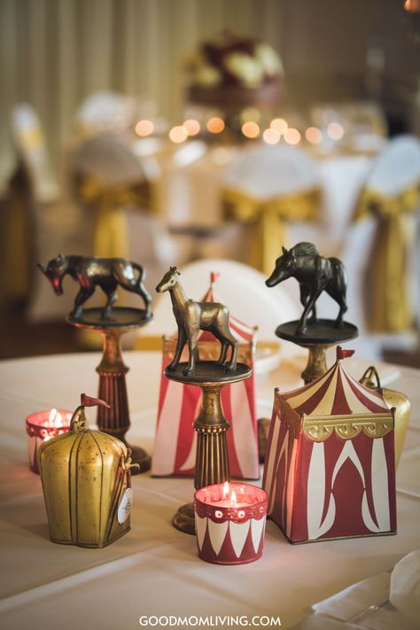 Decorative circus-themed table centerpiece featuring vintage animal figurines and red and white striped tents, set against a backdrop of elegantly draped chairs with golden sashes and soft candlelight. Vintage Carnival Birthday Party, Circus Theme Dinner Party, Big Top Circus Party, Carnival Home Decor, Circus Table Setting, Carnival Christmas Party Theme, The Greatest Showman Theme, Carnival Table Centerpieces, Circus Centerpieces Ideas