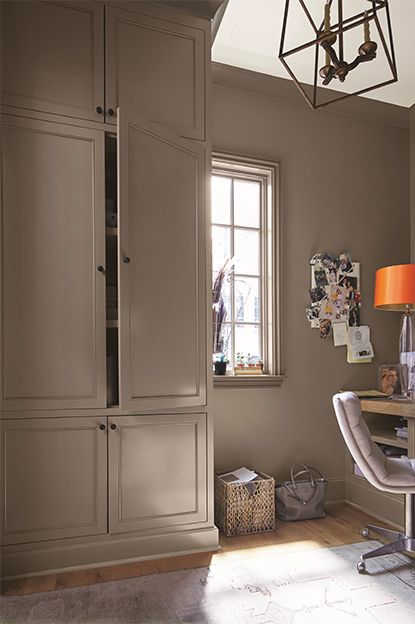 A taupe-painted home office with cupboard, beige chair, wooden desk, note board, white ceiling, and abstract chandelier. Best Dark Beige Paint Color, Dark Beige Kitchen Walls, Dellwood Sand Benjamin Moore, Built In Bookcase Color Ideas, Bromley Taupe Paint, Rich Taupe Paint Color, Falcon Brown Benjamin Moore, Taupe Walls And Trim, Taupe Wall Paint Colors