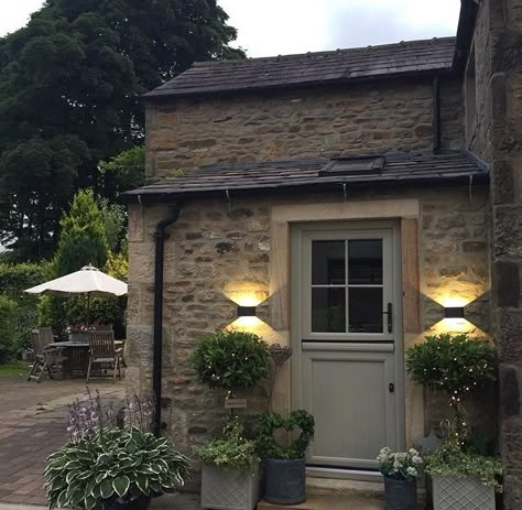 Small stone porch with a green door. The down lighting and little fairy lights in the trees make this a very special entrance. A few garden tubs add to an attractive area in a small space. Stone Porch Ideas Farmhouse, Green Door Cottage, Exterior Cottage Lighting, Stone Porch Ideas Entrance, Outdoor Porch Lights Front Doors, Door Plants Entrance, Front Door Stone House, Light Stone House Exterior, Cottage Doors Front Entrance