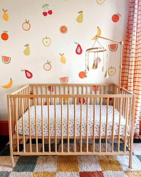 Macrame mobile above wood crib in nursery with fruit painted on wall. Colorful Unisex Nursery, Nursery Fruit Theme, Painting Nursery Walls, Fruit Nursery Decor, Food Themed Nursery, Fruit Themed Bedroom, Colorful Baby Girl Nursery, Girl Nursery Colorful, Painted Nursery Walls