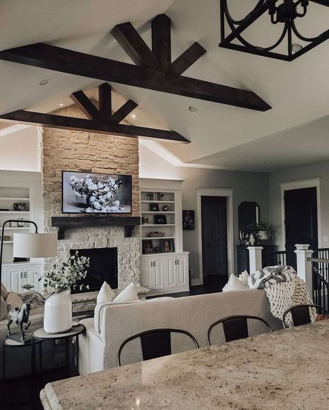 Black stained wood ceiling trusses in living room are mounted over a beige couch. The couch sits opposite a white to tan gradient painted stone fireplace with black wood mantel. Wood Beam Ceiling Living Room, Beams In Living Room, Stained Wood Ceiling, Ceiling Trusses, Ceiling Beams Living Room, Stained Wood Beams, Painted Stone Fireplace, Black Stained Wood, Beams Living Room