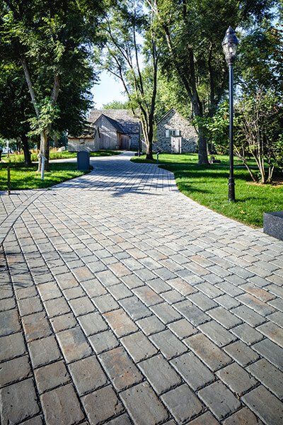 This driveway design is inspired by our Pure paver. Pure brings the larger cobblestone look to permeable pavements for residential and commercial driveways and patios. This interlocking permeable pavement was designed to allow for the proper percolation of surface water into the ground; reducing the risk and severity of flooding, eliminating puddles and speeding up the melting process of snow. Check out our website to shop the look! Side Of Driveway Landscaping, Grey Driveway, Driveway Pavers Design, Driveway Landscaping Ideas, Entrance Landscaping, Driveway Entrance Landscaping, Cobblestone Driveway, Permeable Pavers, Patio Pavers Design