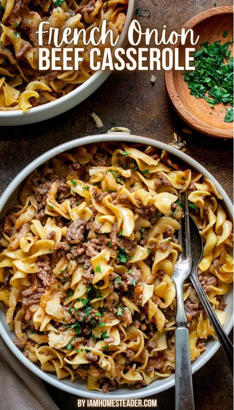 A beige bowl is filled with French onion beef casserole and has a silver spoon and fork in it. Around the bowl is a small wooden bowl of parsley, and another identical bowl of French onion beef casserole, all on a dark countertop. French Onion Beef And Noodles Casserole, French Onion And Beef Noodles, French Onion Beef Noodle Bake 12 Tomatoes, Creamy French Onion Beef And Noodles, French Onion Soup Hamburger Recipes, Ground Beef And French Onion Soup, What To Make With French Onion Soup, French Onion Beef Casserole Recipes, French Onion Noodle Casserole