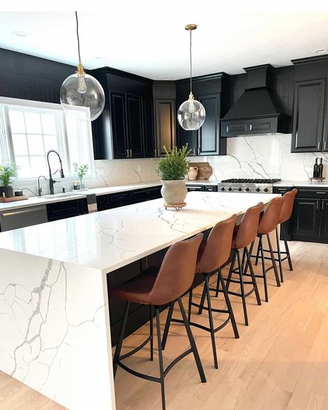 Black cabinets create a life-changing atmosphere in this kitchen, allowing the ribbons of gray to stand out more vibrantly in a white quartz backsplash. If you are reluctant to include another color in your kitchen, glass globe pendant lights can illuminate your space without adding a potential color clash. An expansive quartz waterfall island is accessorized with comfy brown counter stools. Calacatta Laza, Modern Kitchen Trends, Waterfall Island Kitchen, Top Kitchen Trends, 2024 Kitchen, Lake House Kitchen, All White Kitchen, Counter Design, Big Kitchen