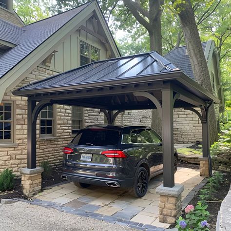 Elegant carport featuring a dark gray metal roof, beautifully complemented by tan stone accents and limestone piers. This open-sided structure shelters a black Volkswagen Tiguan, set against an old cottage-style house with black windows. The landscaping boasts lush greenery, colorful flowers, and two large decorative brown blocks enhancing the aesthetic. Carport Beside House, Beautiful Carport Ideas, Carport Driveway Ideas, Front Of House Carport, Unattached Carport Ideas, Pergola As Carport, Cottage Carport Ideas, Detached Carport Ideas Breezeway, Carport With Privacy Wall