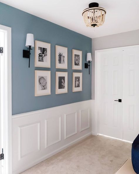 This traditional space features a stormy blue-painted wall and white wainscotting. A beaded light fixture hangs from the ceiling. The wall displays six rectangular frames with light wood frames around black and white family photos. Two traditional black scones with white shades are mounted on either side of the photos. Wall Decor For Blue Walls, Light Blue Paneled Walls, Light Blue Hallway Walls, Black Ceiling Light Fixtures Bedroom, Blue And White Hallway Ideas, Blue And Cream Hallway, Living Room Beading Ideas, Wall Half Wood Half Paint, Light Blue Entryway Walls