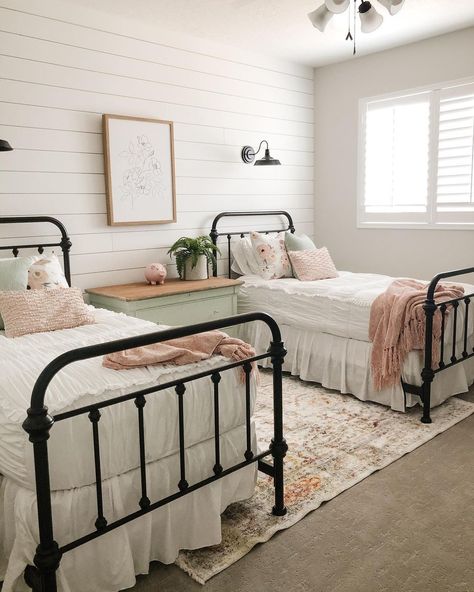 Twin black metal spindle beds are dressed in white and pale pink bedding. A two-toned stained wood and light green dresser stands between the beds in front of a white shiplap feature wall. Sisters Bedroom Ideas, Twin Beds Guest Room, Twin Girl Bedrooms, Sister Bedroom, White Metal Bed, Shared Girls Room, Sister Room, Shared Girls Bedroom, Big Girl Bedrooms