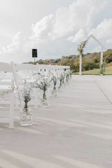 Ceremony floral installation on white arbor with textural white and neutral flowers and greenery. Clear glass jugs with greenery lining the ceremony aisle Wedding Isles Decoration Chapel, Glass Jugs Wedding Aisle, Eucalyptus Down The Aisle, Isle Ceremony Decorations, White Flowers Lining Wedding Aisle, Flower Vases Down Aisle, Neutral Ceremony Decor, Wedding Aisle Flower Vases, Glass Jug Wedding Aisle