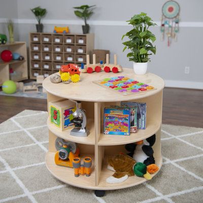 The circular storage from wood designs gives parents and teachers a place to stow their kids’ belongings in an angle-free fashion. The natural aesthetic provides simple curved elegance for any classroom, daycare, or home environment. This table’s 2-tiered design adds ample storage options for toys, crafts, and books. You can quickly change the flow of the room by moving this table easily with its locking casters. Size: 23.5" H x 29.5" W x 29.5" D | Wood Designs Circular Double Sided Shelving Uni Modern Daycare Design, Daycare Interior Design, Circular Storage, Preschool Storage, Daycare Design Ideas, Montessori Design, Classroom Architecture, Childcare Rooms, Wood Shelving Units