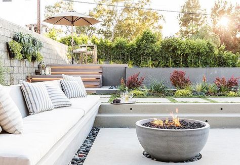 Sunken patio is filled with a concrete bench lined with white outdoor cushions and gray striped outdoor pillows faces a concrete bowl fire pit surrounded by black river rocks. Sunken Patio, Wood Burners, Rooftop Terrace Design, Fire Pit Seating, Pergola Design, Modern Backyard, Steel Wood, Beach House Interior, Terrace Design