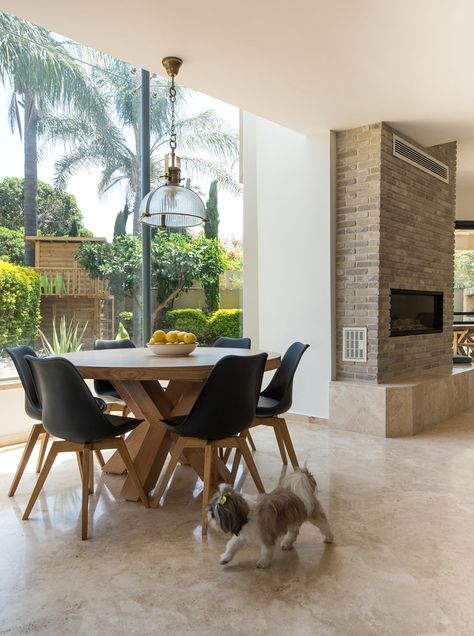 A white round table with black chairs, in front of a fireplace and glass walls with a view to the garden outside, with a little dog wondering by. By Liat Hadas, Architecture & Design. Round Table With Black Chairs, Table In Front Of Fireplace, Table With Black Chairs, Architecture Table, Curtain Styling, White Round Table, Table Fabric, White Round Tables, Contemporary Mediterranean