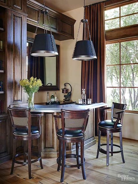 Extending out from a windowed corner, this handsome home bar takes its cue from the room's British-study leanings and paneled woodwork. A back-bar mirror, strikingly shaded pendant lights, and a white marble bar top draw attention to the drink station, which is outfitted with cozy leather seating. Home Bar Window, Bar Pass Through, Corner Bars For Home, Home Bar With Seating, Bar With Window, Corner Wet Bar, Corner Bar Ideas, Corner Home Bar, Basement Pub