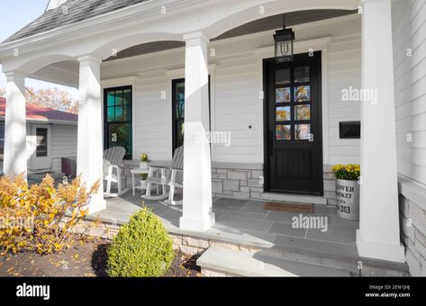 Download this stock image: A new, white modern farmhouse with a dark wood door with windows, white pillars, a stone floor, and patio furniture. - 2EN1JHJ from Alamy's library of millions of high resolution stock photos, illustrations and vectors. Colonial Porch Ideas, Porch Columns Makeover, House Pillar Design, Summer Welcome Sign, House Pillars, Welcome Sign Porch, Front Porch Remodel, Front Door Welcome Sign, Door Welcome Sign