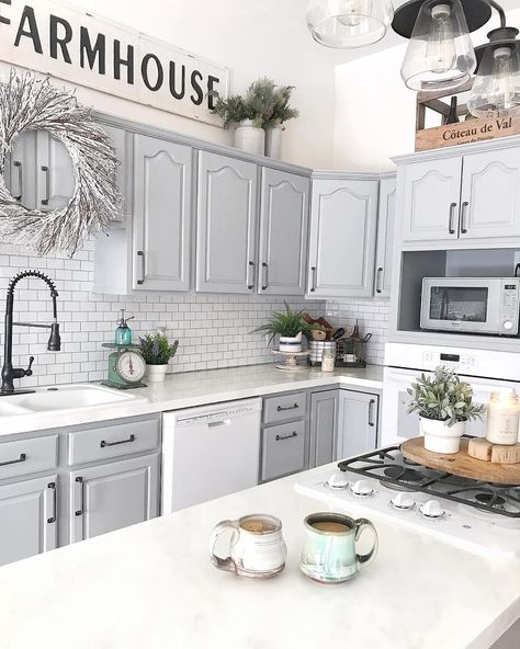 This kitchen features white walls, light grey cabinets with black handles, and white counters. Potted plants in white pots are strewn about the kitchen in front of the white subway tile backsplash with a light grey grout. Grey Cabinets White Countertops, Cabinets With Black Handles, Light Grey Cabinets, White Kitchen Counters, Gray Grout, Light Grey Kitchen Cabinets, Grey Kitchen Walls, Grey Kitchen Floor, Light Grey Kitchens