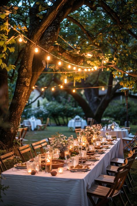 Outdoor table setup with white cloth, candles, and flower centerpieces under string lights hanging from tree branches in a garden during twilight. Garden Celebration Party Ideas, Backyard Autumn Wedding, Dinner Candles Decoration, Backyard Garden Dinner Party, Outdoor Garden Party Ideas Decoration, Elegant Backyard Dinner Party, Summer Garden Dinner Party, Engagement Party Garden, Backyard 40th Birthday Party