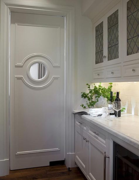 A swing door accented with decorative trim moldings and a round window opens to a butler's pantry boasting leaded glass upper cabinets stacked above drawers over white inset cabinets paired with white marble countertops and a white subway tiled backsplash. Glass Upper Cabinets, Kitchen Butlers Pantry, Glass Pantry Door, Butler’s Pantry, White Subway Tile Backsplash, Swinging Doors, White Marble Countertops, Butler Pantry, 아파트 인테리어