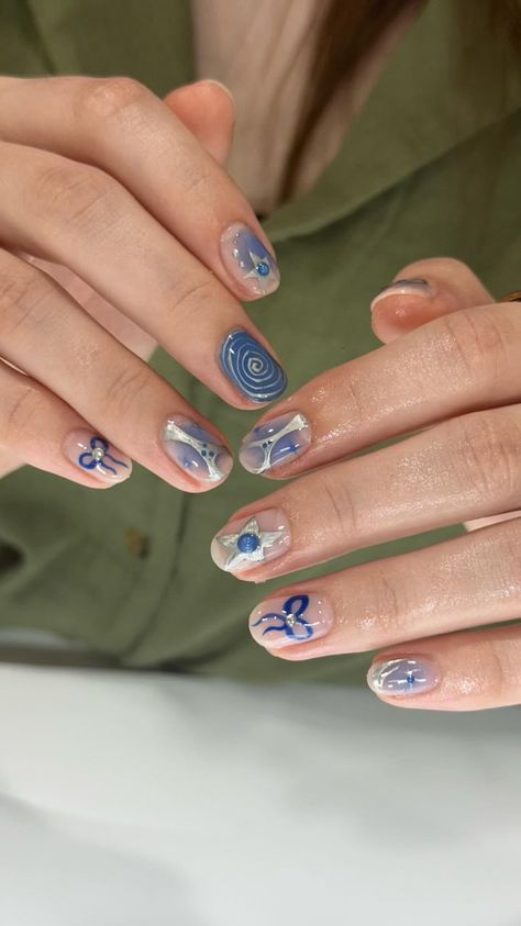 The image shows a close-up of a person's hands with an intricate blue-themed gel nail art. The designs include various patterns such as spirals, abstract shapes, stars, and bows, all in shades of blue and silver. The background is a light olive green shirt worn by the person, creating a contrasting backdrop for the nail designs. The nails have a glossy finish, highlighting the detailed and artistic work. The hands are well-manicured, and the skin appears smooth. Trendy Blue Nails Short, Stars Nails Short, Blue Mismatched Nails, Short Blue And Silver Nails, Blue And Gray Nail Ideas, Short Nail Designs Easy Simple, Blue And Silver Short Nails, Funky Nails 2024, Silver Nail Designs Short