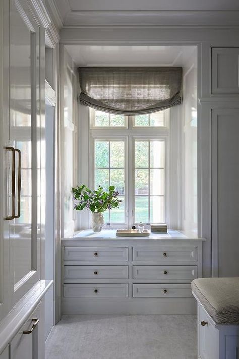 Chic walk in closet features a white built in dresser with brass knobs under a window with tan shade. Closet Between Windows, Built In Dresser Under Window, Built In With Slanted Ceiling, Pass Through Closet To Bathroom, Closet Designs With Window, Built In Linen Closet Hallway, Chic Walk In Closet, Hallway Linen Closet Built Ins, Closet With Built In Dresser