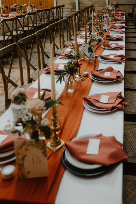 Wedding reception table decor with long burnt orange table runner and burnt orange napkins. Taper candles stand in brass candle holders & elegant Autumn flowers sit in small glass bud vases. Long trestle tables with cross back chairs. Wedding Table With Table Runner, Wedding Table Settings Burnt Orange, Burnt Orange And Brown Wedding Decor, Burnt Orange Table Runner Wedding, Autumn Wedding Diy Decorations, Rustic Wedding Decor Autumn, Burnt Orange Round Table Setting, Fall Wedding Simple Table Decor, Wedding Reception Table Runners