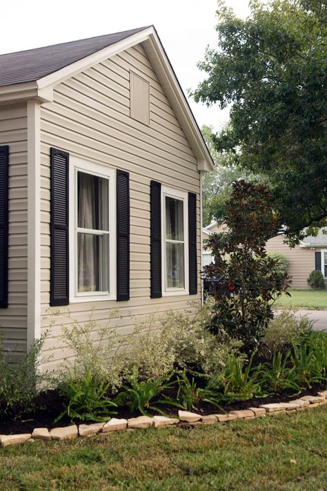 Fixer Upper hosts Chip and Joanna Gaines repainted the siding a neutral shade and the shutters black for a fresh updated to the exterior. They also updated the landscaping and added new grass. Exterior Door Colors With Tan Siding And Black Shutters, Tan Ranch House Exterior, Black Front Door Tan House, Tan And Black Exterior House, Tan Siding House, Tan Siding Black Shutters, Beige House Black Shutters, Tan Exterior House, Tan And Black House Exterior