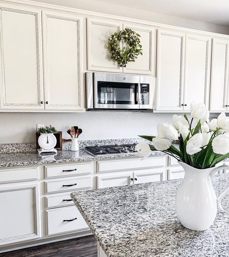 This kitchen features black and white backsplash with granite counters. White cabinets with black hardware encase stainless steel microwave tucked above gas range. Porcelain pitcher with an array of white flowers is set upon countertop. White Cabinets Black And White Granite, Granite Backsplash With Tile Above, Black And White Kitchen Granite, Granite Countertops With White Cabinets Grey Backsplash Ideas, White And Black Granite Countertops, Black And White Granite Kitchen, Black And White Granite Countertops, Tile Backsplash Kitchen Ideas, White Cabinets With Black Hardware