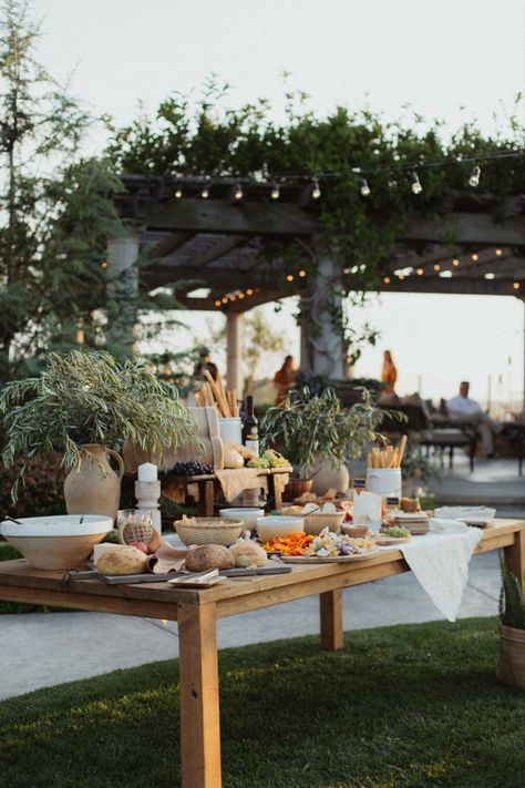 A photo of a large charcuterie table. It has large vases of olive branches and different breads. Bowls filled of seasonal fruits, hummus, and sliced bread. Looks like it is right out of the coast of Italy. Charcuterie Table With Flowers, Charcuterie Station Wedding, Wedding Canape Table, Garden Charcuterie Board, Outdoor Grazing Table, Appetizer Table Wedding, Summer Wedding Buffet, Charcuterie Board Wedding Reception, Engagement Party Food Table