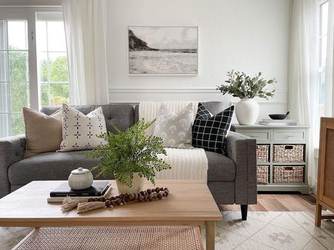 This brightly lit living room showcases a dark grey couch decorated with brown, grey, white, and black throw pillows. A light wood coffee table sits in front of the sofa accessorized with stacked books and a potted plant. A light green side table with woven baskets rests on the side of the sofa. Charcoal Grey Sofa With Cushions, Black And White Throw Pillows Grey Couch, Dark Gray Couch Green Pillows, Grey Couch And Coffee Table, Dark Grey Couch, Grey Sectional Couch, Light Gray Couch, Grey And Brown Living Room, Dark Grey Couches