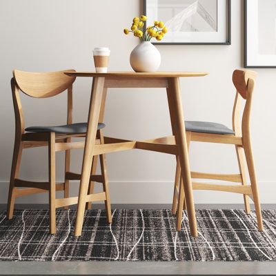 Inspired by iconic mid-century silhouettes, this 3-piece dining set creates a cohesive and minimal look in your kitchen or dining room. It includes a counter-height table and two stools, all crafted from solid and engineered wood with a light, natural oak finish that complements any modern space. The table is built on top of splayed legs with X-shaped support, and its round table top is designed to accommodate two guests after assembly. Both stools showcase curved backrests and built-in footrest Tall Dining Room Table, Small Round Kitchen Table, Small Dinner Table, Small Breakfast Table, Small Round Dining Table, Round Bar Table, Apartment Dining Room, Round Kitchen Table, Small Kitchen Tables