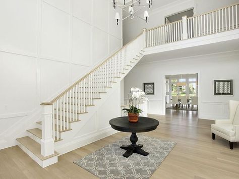 Two story foyer features a Jonathan Adler Ventana 2 Tier Chandelier illuminating a round black entry table atop a gray Moroccan tile rug placed next to a staircase with board and batten walls. Black Entry Table, Double Height Entrance, Cottage Foyer, Round Foyer, Round Foyer Table, White Staircase, Batten Wall, 2 Story Foyer, Traditional Staircase