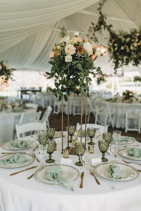 A gold floral vessel, a pink, peach and white arrangement sits at the top, below is gold trimmed plates and green goblets with matching gold silverware. Green Wedding Theme Receptions, Blush Sage And Gold Wedding Decor, Emerald Green Wedding Place Settings, Olive Green Reception Decor, Emerald Green And Gold Wedding Ideas, Olive Green Wedding Decorations, Sage Green And Gold Wedding Table Decor, Sage Green Reception Decor, Olive Green Wedding Table Decor