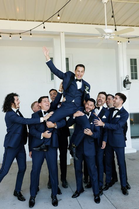 The couple wanted the wedding party to complement the environment of the venue in a timeless and upscale way that reflected the blue lake and beautiful surrounding greenery. The bridesmaids wore dusty blue Azazie dresses with soft, natural glam by LJ Artistry and Reborn Hair Company, and carried bouquets full of soft green, soft pinks, and creams. The groomsmen wore navy blue suits with black lapels and bowties from Men's Wearhouse. #lakewedding #groomsmen Wedding Groomsmen Attire Navy Blue, Cobalt Blue Groomsmen Attire, Blue Tuxes Wedding, Dark Blue Summer Wedding, Navy And Black Tux Wedding, Navy And Dusty Blue Groomsmen, Groomsmen Blue Tie, Navy Blue Bridesmaids And Groomsmen, Dark Navy Blue Groom Suit